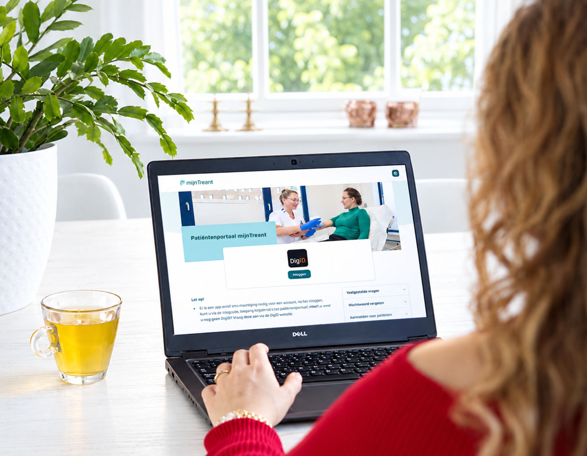 Vrouw gebruikt een laptop aan een tafel naast een kopje thee en een plant.